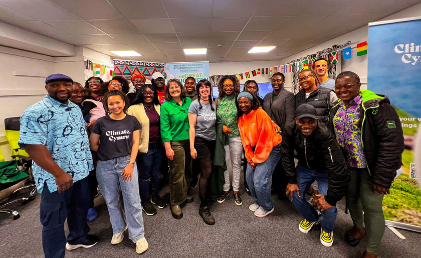 A diverse group of people stands smiling together in a decorated room, with colourful flags hanging on the walls and Climate Cymru banners visible in the background.