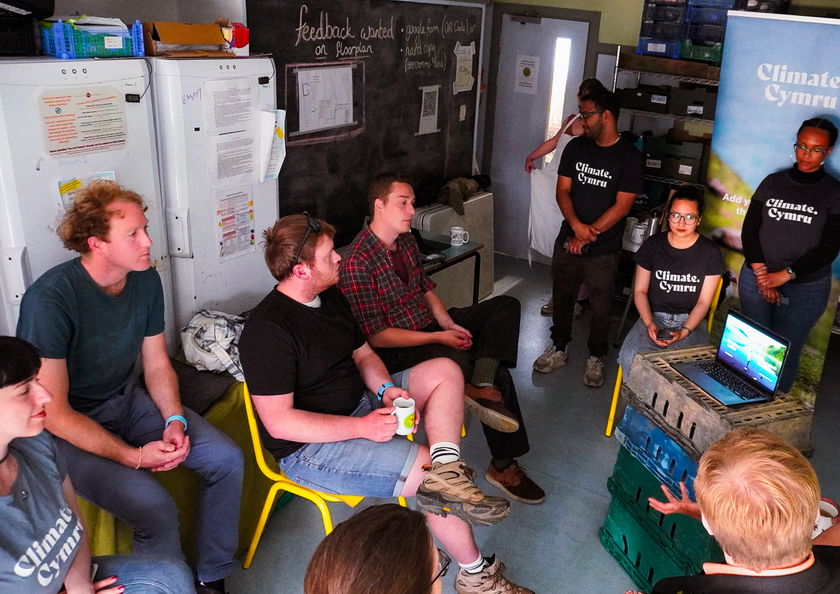 A group of people sit and stand in a small room, attentively watching a young person present with a laptop. Several wear Climate Cymru shirts, and informational posters are visible on the walls.