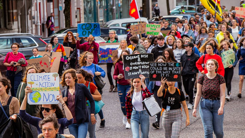 A large group of people march down a city street holding colourful signs about climate action and environmental protection during a protest or rally. The crowd appears diverse and determined.