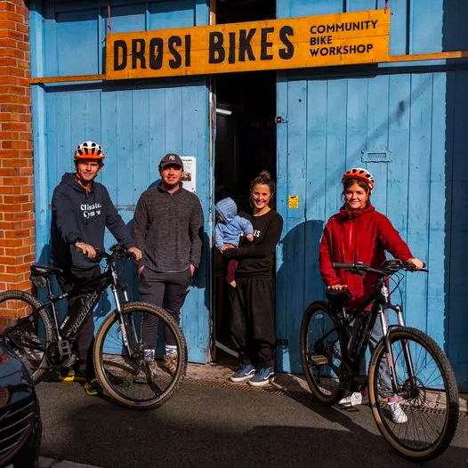 Four people, two with bikes and helmets, stand outside a blue-painted building with a yellow sign reading DROSI BIKES Community Bike Workshop. Two others, one holding a child, stand in the doorway.