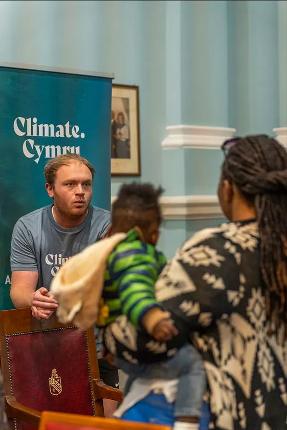 A man in a Climate Cymru shirt sits behind a table, talking to a woman holding a baby. They are indoors, and a blue Climate Cymru banner is visible in the background.