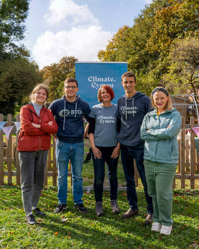 Five people stand together outdoors on grass, smiling at the camera. They are in front of a wooden fence and a blue Climate Cymru sign. Trees and bunting are visible in the background on a sunny day.