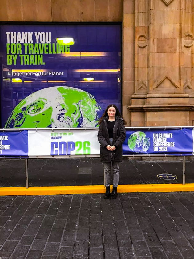 A young woman stands in front of a large COP26 poster and a sign reading Thank you for travelling by train with a stylised Earth graphic, outside a building with stone columns.