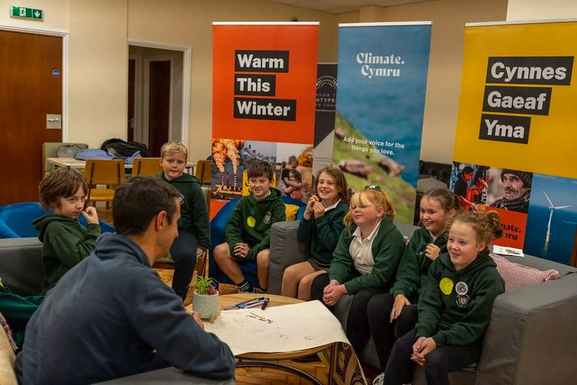 A group of children in green hoodies sit around a table, listening to an adult in a casual room. Colourful banners behind them read “Warm This Winter,” “Climate Cymru,” and “Cynnes Gaeaf Yma.”.