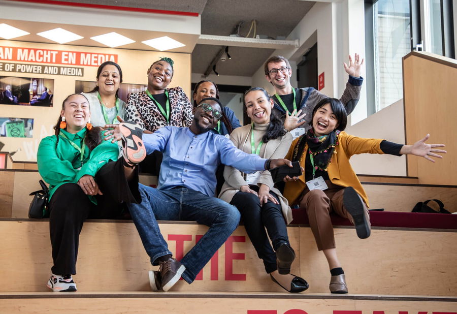 A group of eight diverse people sit on bleachers indoors, smiling and joyfully posing with arms outstretched. The setting appears to be modern and bright, with media-related signage visible in the background.
