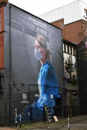 A large mural on a brick building depicts a woman in blue scrubs and a face mask, titled “MELANIE, MARCH 2020.” A ladder leans against the wall below the mural, which is part of the National Portrait Gallery’s “Hold Still” project.