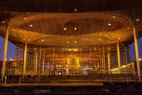 The image shows the Senedd building in Cardiff, Wales, illuminated at night. Its modern wooden roof curves above glass walls, with warm lighting highlighting the architectural details and broad entrance steps.