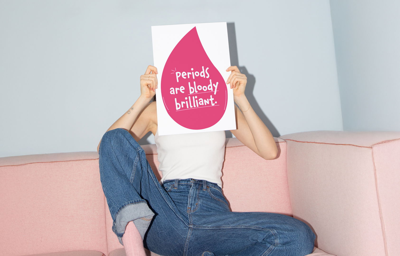 A person sitting on a light pink sofa holds a bright red poster in front of their face. The poster reads: Let’s talk about... Bloody Brilliant Periods! and provides details for joining the conversation.