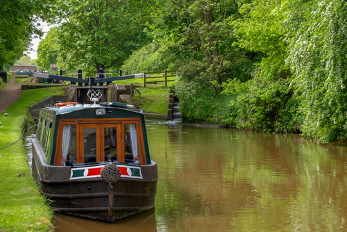 A narrowboat is moored on a calm canal surrounded by lush green trees and grass, with a lock and small waterfall visible in the background on a peaceful, overcast day.