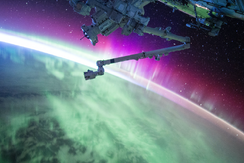 A view from the International Space Station shows vibrant green and pink auroras glowing over Earth, with part of the station’s robotic arm visible against the backdrop of space and stars.