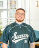 A smiling young man with glasses and a beard wears a dark green Seaszn 19 baseball jersey. He stands indoors in a bright room with glass walls, plants, and a blue chair in the background.