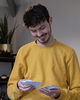 A smiling person in a yellow jumper looks down while holding several small cards or papers in their hands. There is a potted plant and shelving in the background.