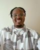 A man with glasses and short plaited hair smiles at the camera. He is wearing a white shirt with black geometric patterns and standing in front of a plain light-coloured wall.