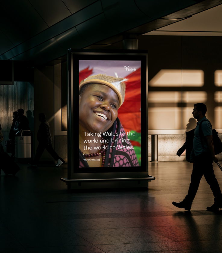 A smiling person in traditional attire is displayed on a digital billboard in an indoor public space. The billboard reads, Taking Wales to the world and bringing the world to Wales. People walk by in the background.