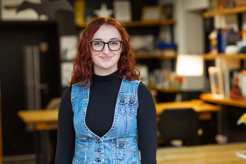 A young woman with red hair and glasses smiles whilst standing indoors. She is wearing a black polo neck and a sleeveless denim waistcoat. The background shows a blurred office or library setting with shelves and tables.