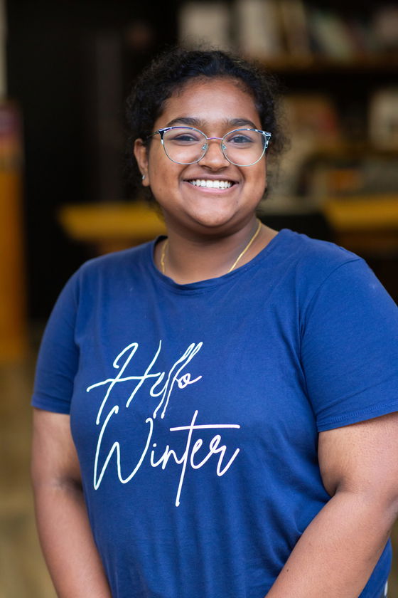 A smiling person with curly hair, glasses, and a blue “Hello Winter” T-shirt stands indoors with blurred shelves in the background.