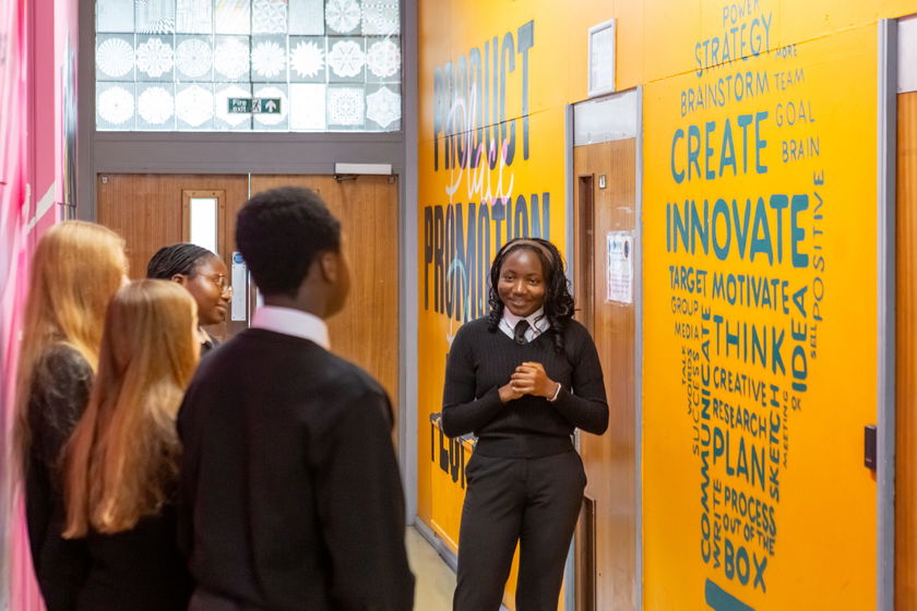 A group of students in school uniforms stand in a corridor decorated with motivational words like Create and Innovate on a bright orange wall. One student is speaking whilst others listen attentively.