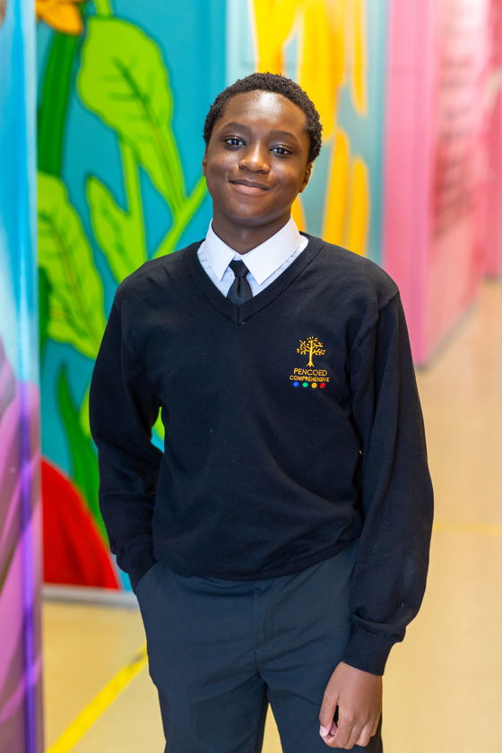 A student wearing a black jumper with a school logo, white shirt, and tie stands smiling in a colourful corridor with bright mural artwork on the walls.