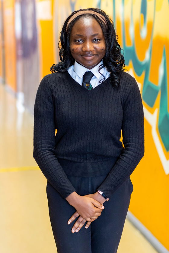 A young woman stands indoors against a colourful orange and green wall, smiling. She wears a black jumper over a collared shirt and tie, with her hands gently clasped in front of her.