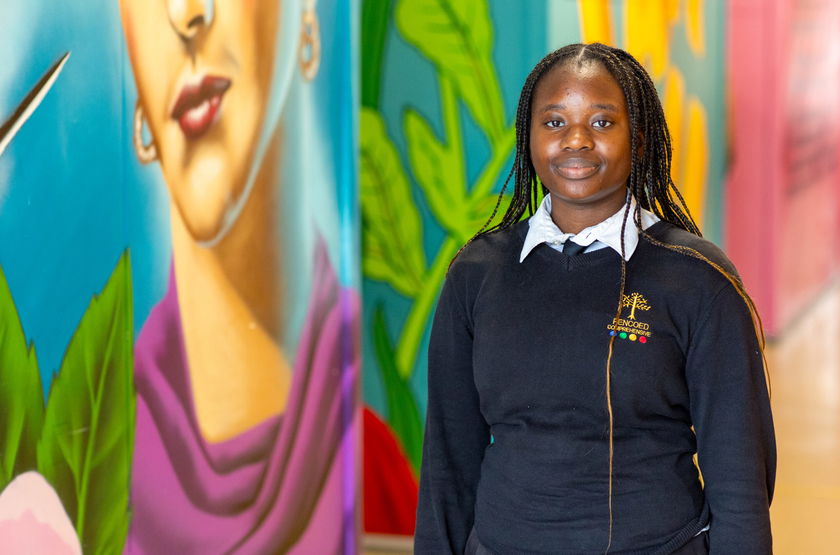 A young woman with plaited hair, wearing a black school jumper, stands smiling in a corridor with colourful mural art on the walls behind her.