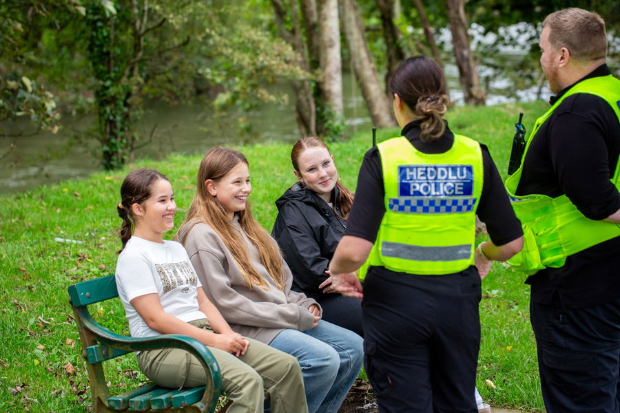 Two police officers speak with three smiling girls sitting on a green bench in a park. The officers wear “Heddlu Police” uniforms. Trees and grass surround the group, creating a relaxed, outdoor setting.
