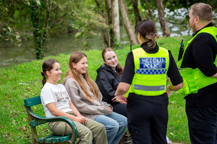 Two police officers speak with three smiling girls sitting on a green bench in a park. The officers wear “Heddlu Police” uniforms. Trees and grass surround the group, creating a relaxed, outdoor setting.