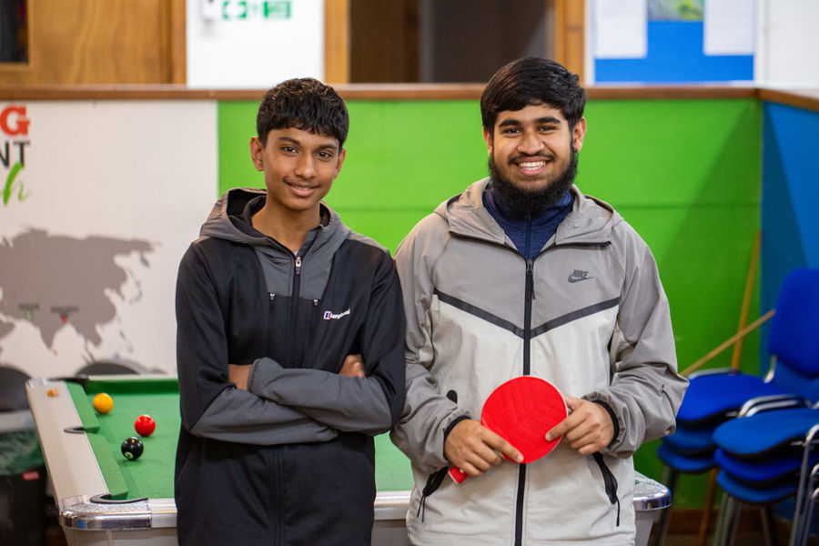 Two young men stand by a pool table; one smiles holding a red table tennis bat, while the other stands with arms crossed. Both wear jackets. The background shows green and blue walls and stacked chairs.