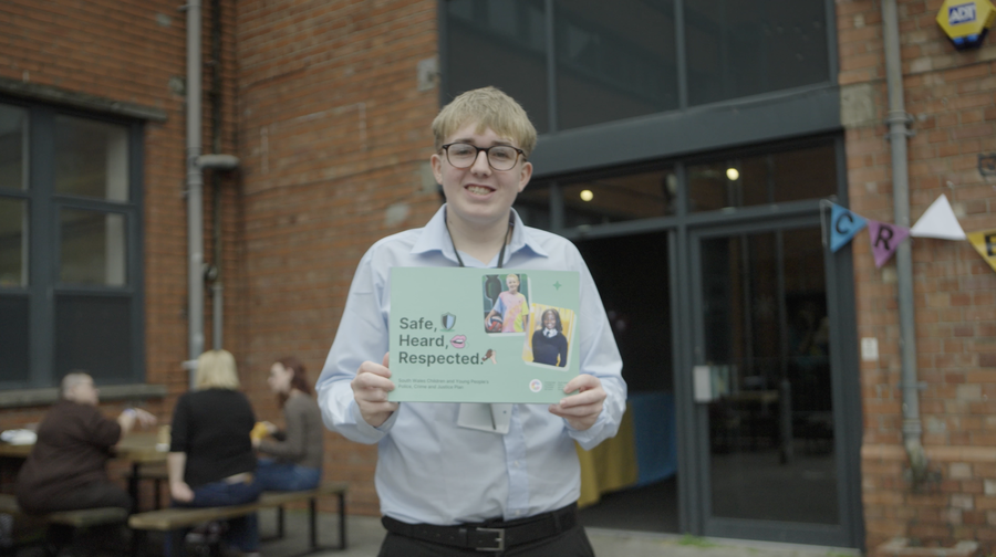A young person wearing glasses and a light blue shirt stands outside a building, smiling and holding a sign that reads “Safe, Heard, Respected” with photos and text on it. People are seated at tables in the background.