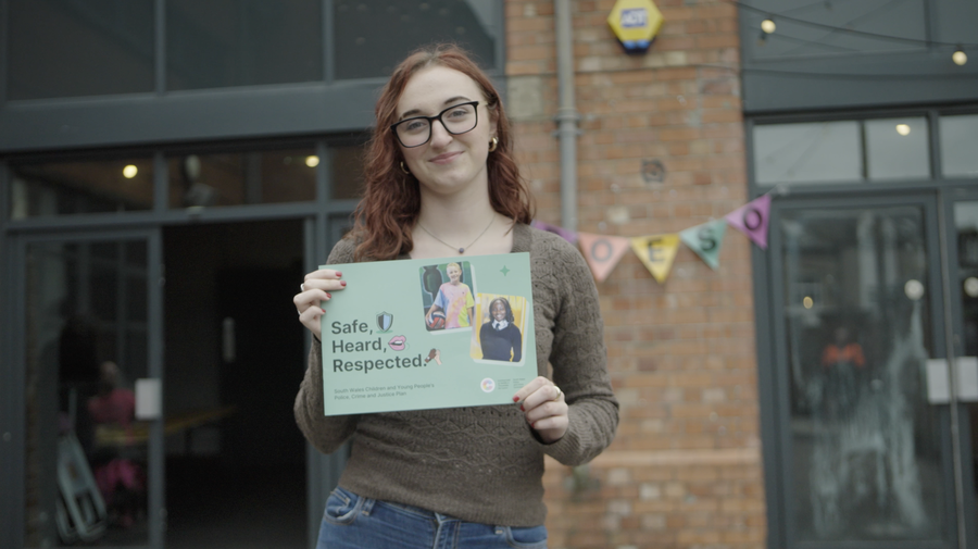 A person with glasses and reddish hair stands outside a brick building, holding a sign that reads Safe, Heard, Respected with photos and colourful designs. There is bunting in the background.