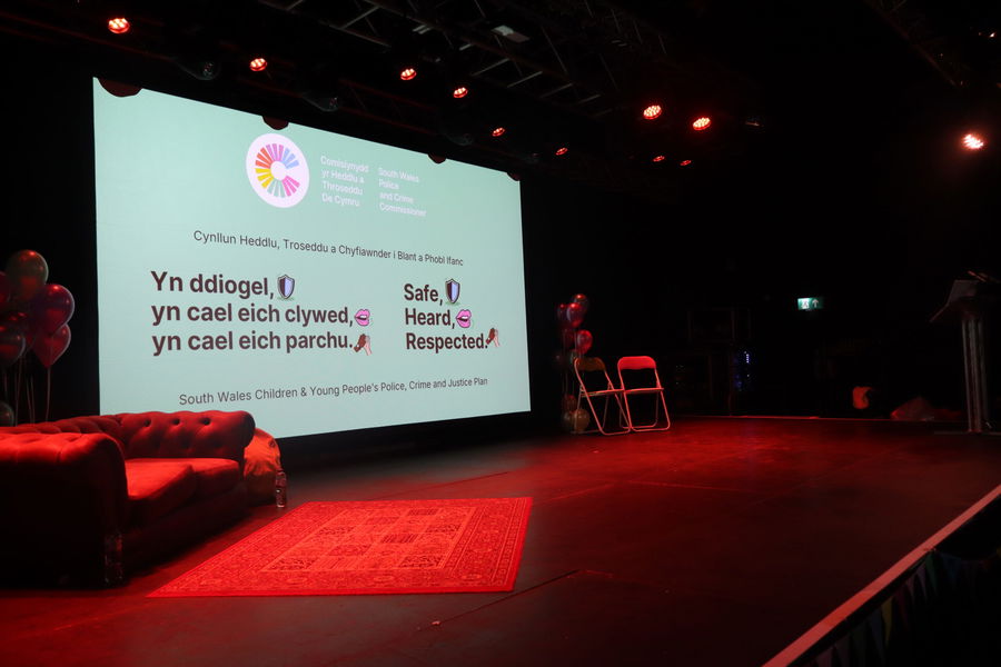 A dimly lit stage with a red sofa, balloons, two chairs, and a lectern. A large screen displays bilingual text about safety, respect, and being heard, with colourful graphics and icons.