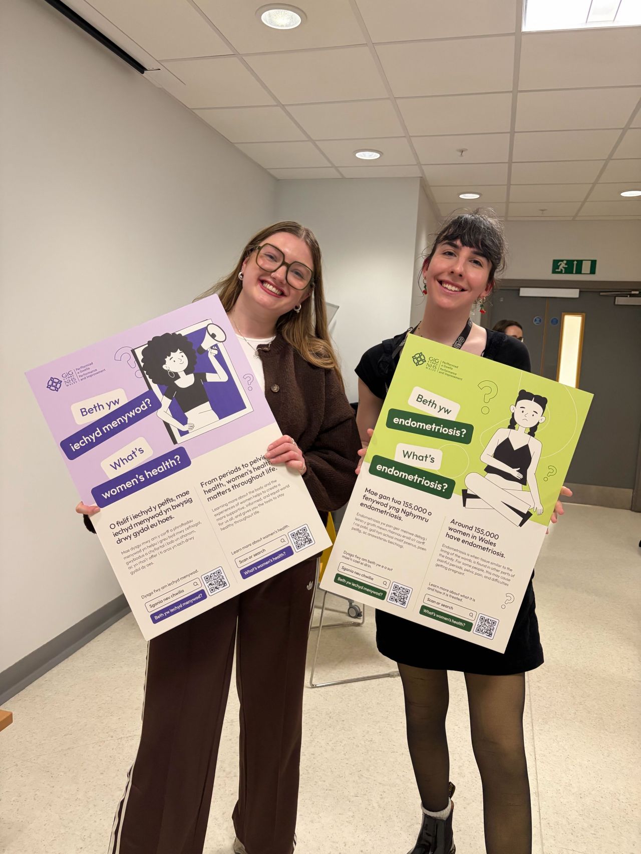 Two women stand indoors, smiling and holding informative 