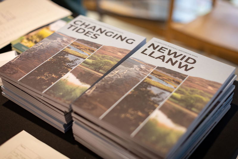 A stack of brochures titled Changing Tides and Newid Llanw, featuring landscape photos of water and land, displayed on a table.