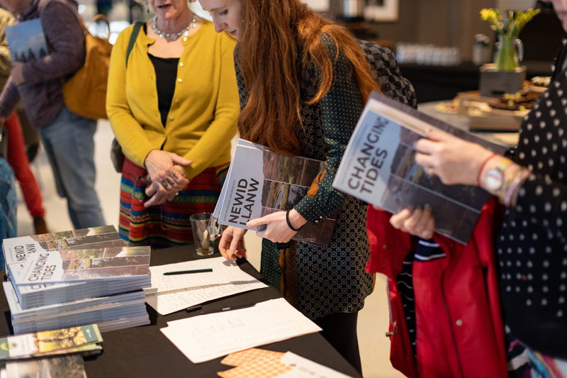 People stand around a table with stacks of books titled Changing Tides and Newid Llanw, signing papers and collecting copies at what appears to be a book launch or registration event.