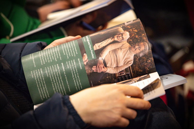 A person holds and reads a magazine or booklet featuring a photo of two older adults smiling and sitting together, with printed text on the page. The person's hands and part of their sleeve are visible.