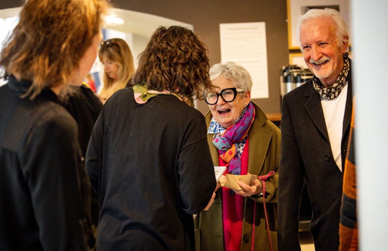 A group of people indoors are engaged in conversation. An older woman wearing glasses and a green coat is smiling and holding a walking stick, while an older man beside her also smiles. Other people are visible in the background.