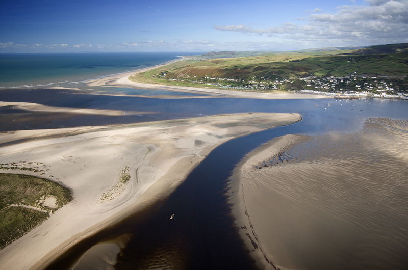 Aerial view of a winding river flowing into the sea, with sandy beaches, tidal flats, and a coastal town set against green hills under a blue sky with scattered clouds.