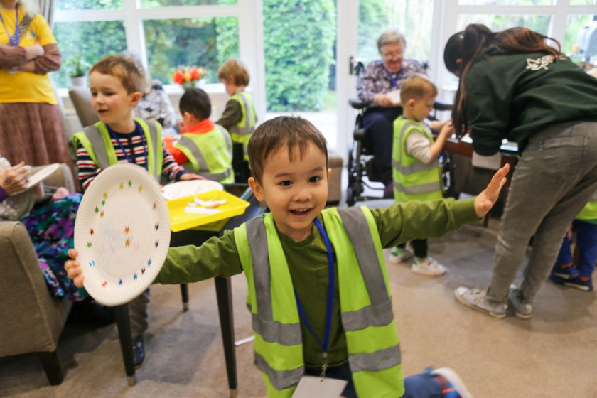 A young child wearing a green safety vest smiles and holds a decorated paper plate during a Glow For Good event in a room with other children and elderly adults, suggesting a joyful group activity or intergenerational gathering.