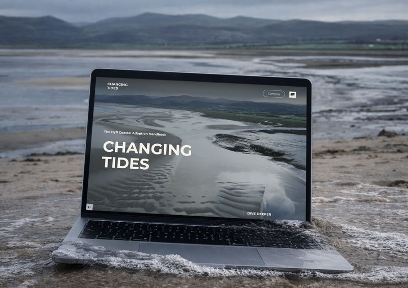 A laptop on wet sand near the water’s edge displays a website titled “CHANGING TIDES”, featuring an aerial photo of a coastal landscape with winding water channels. Hills and cloudy skies are visible in the background.