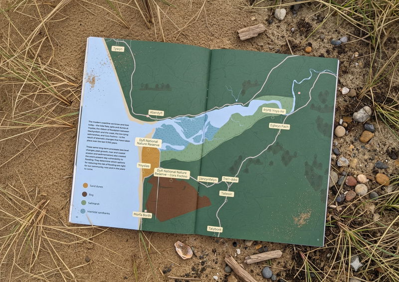 An open booklet showing a colourful map of a coastal region, surrounded by sand, pebbles, and dry grass. The map highlights rivers, marshes, nature reserves, and several labelled locations.