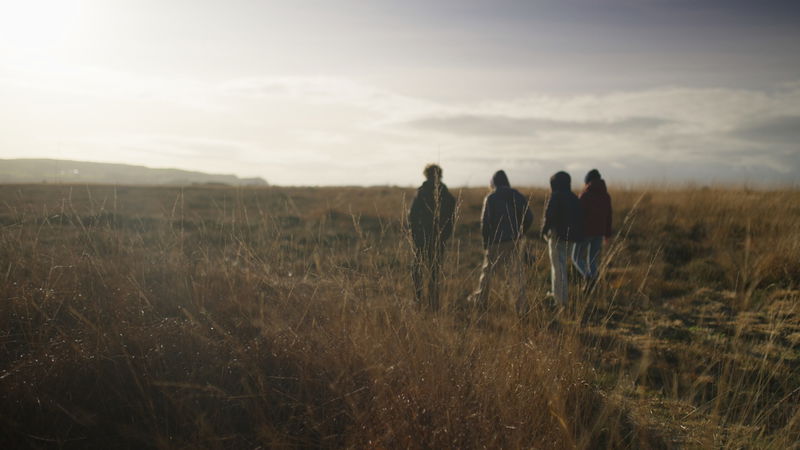 Four people walk through a field of tall grass under a cloudy sky at sunset, with their figures silhouetted against the light in the distance.
